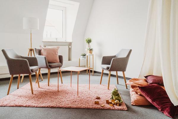 Therapieraum mit drei Stühlen, einem Tisch, Pflanzen, einem rosa Teppich, Spielsachen und einer Sitzecke