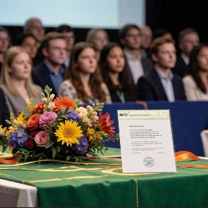 Preisverleihungszeremonie mit Publikum, Blumenarrangement und Urkunde - KI generiert