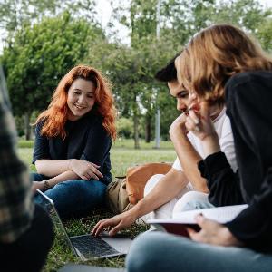 Im Gras sitzende junge Menschen, die gemeinsam in einen Laptop schauen.