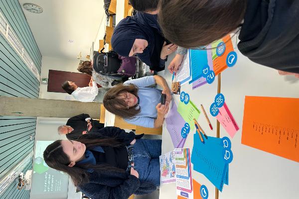 Students gathered around a table examining coloured cards with linguistic landscape taxonomies and workshop materials. The image illustrates collaborative analysis, discussion, and hands-on engagement with methods of linguistic landscape research.