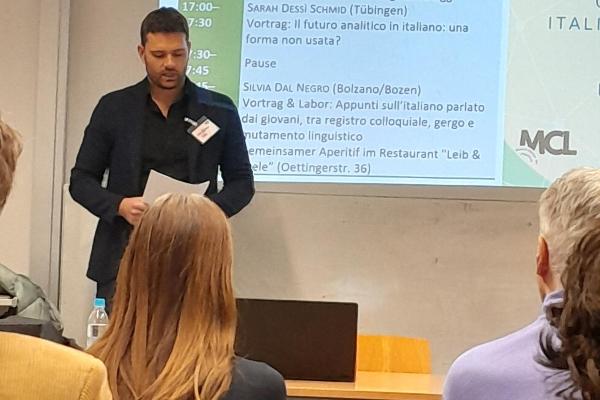 Von hinten fotografiert die Zuhörerschaft und eine vortragende Person (evtl Mann mit Papier in der Hand) mit Beamer-Projektion