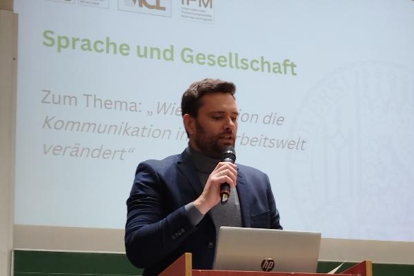 Mann mit Mikro vor Rednerpult im Hintergrund Beamer-Projektion mit Aufschrift Sprache und Gesellschaft