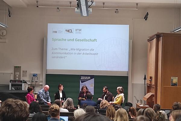 Von hinten im Hörsaal fotografierte, weiter entferne Bühne mit DiskutantInnen über die Köpfe der Zuhörenden hinweg