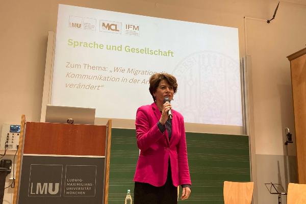 Frau rechts neben Rednerpult stehend in der rechten Hand ein Mikro im Hintergrund Beamer-Projektion mit Aufschrift Sprache und Gesellschaft