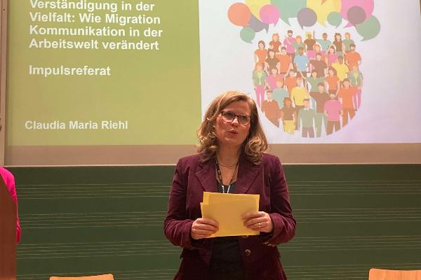 Frau auf Bühne stehend mit Papieren in der Hand im Hintergrund Beamer-Projektion mit Aufschrift Wie Migration unsere Arbeitswelt beeinflusst