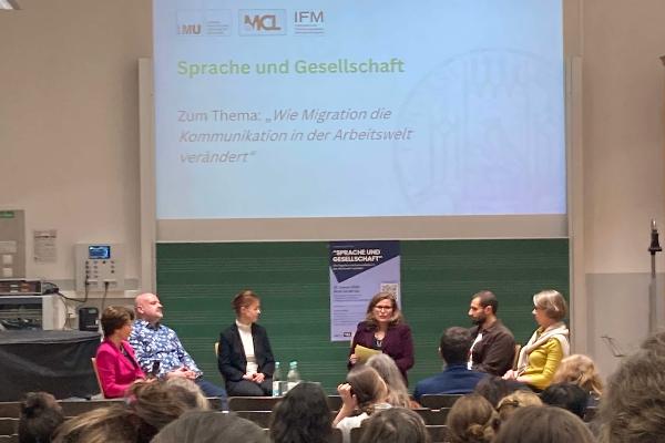 Von hinten im Hörsaal fotografierte, weiter entferne Bühne mit DiskutantInnen über die Köpfe der Zuhörenden hinweg