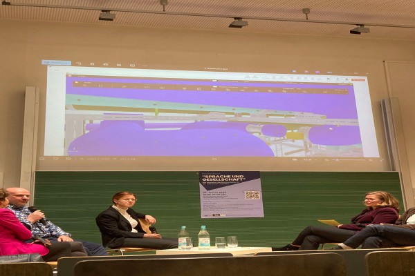 Nahaufnahme der Bühne auf drei sitzende Personen, die diskutieren,eine Person hält ein Mikro, im Hintergrund ist eine Beamer Projektion eines Gebäude Entwurfes