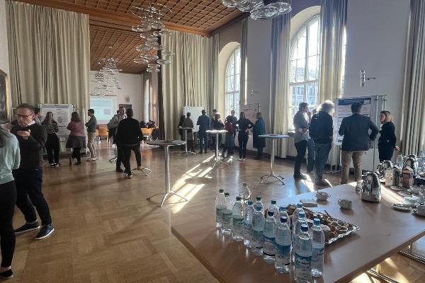 Saal mit einem Tisch mit Gläsern, Tassen, Kannen und Essen, Personen stehen im Saal an Stehtischen