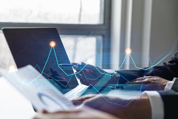 A laptop with a digital chart overlay in blue and purple showing upward and downward data curves, held by someone typing | © iStock A laptop with a digital chart overlay in blue and purple showing upward and downward data curves, held by someone typing