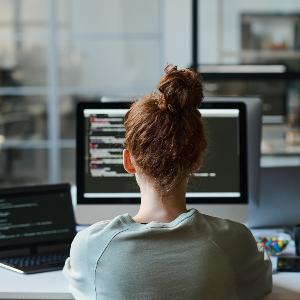 Woman working with code on a computer with multiple screens in a modern office setting.