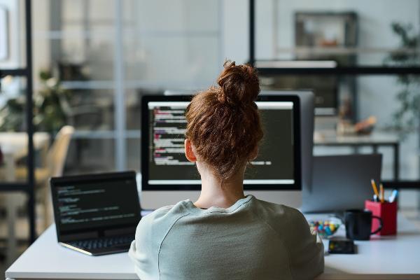 Woman working with code on a computer with multiple screens in a modern office setting.