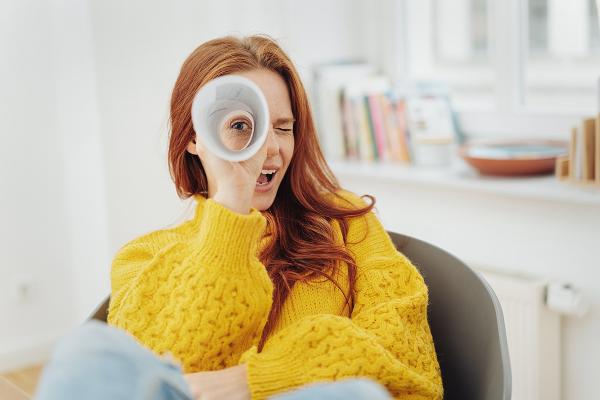 Symbolbild: Eine junge Frau schaut mit einem Auge durch ein zusammengerolltes Blatt Papier.
