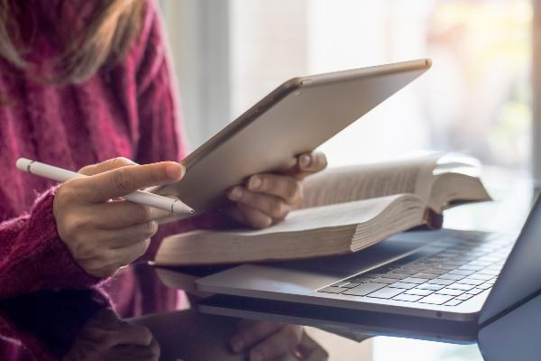 Symbolbild: Eine Person hält einen Laptop und einen Stift, auf dem Tisch liegen ein Buch und ein Laptop.