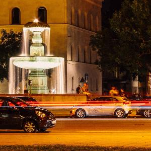 Die Ludwigsstraße bei Nacht, auf welcher Autos am beleuchteten Brunnen der LMU vorbeifahren.