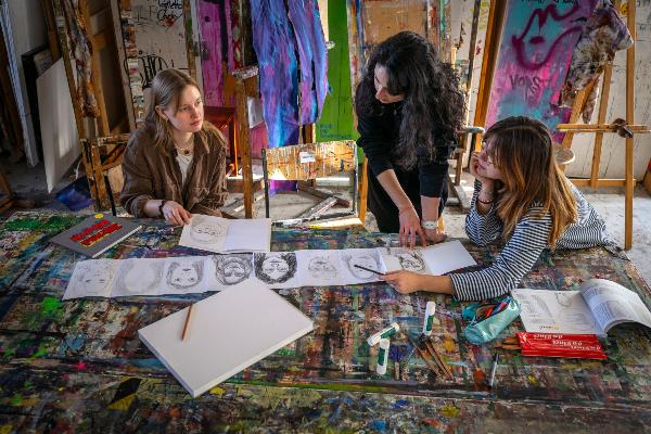Three students look at drawings. In the background you can see many colorful works of art and easels.