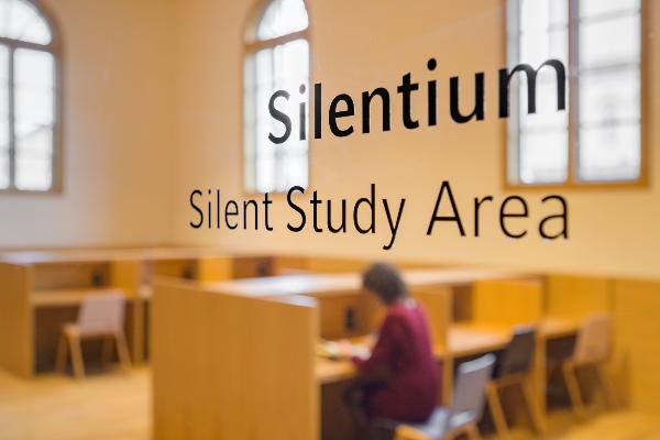 Blick in einen Raum des Philologikums durch eine Glaßtür mit der Aufschrift Silentium, in dem eine Studentin liest.