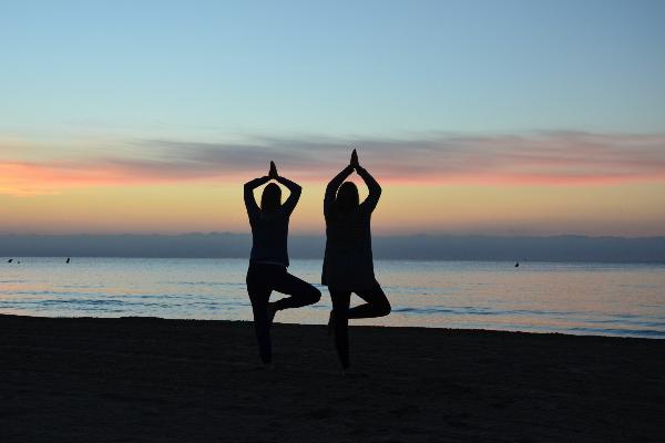 Zwei Studentinnen machen eine Yogafigur an einem Strand vor einem Sonnenuntergang.