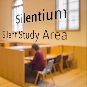 Blick in einen Raum des Philologikums durch eine Glaßtür mit der Aufschrift Silentium, in dem eine Studentin liest.