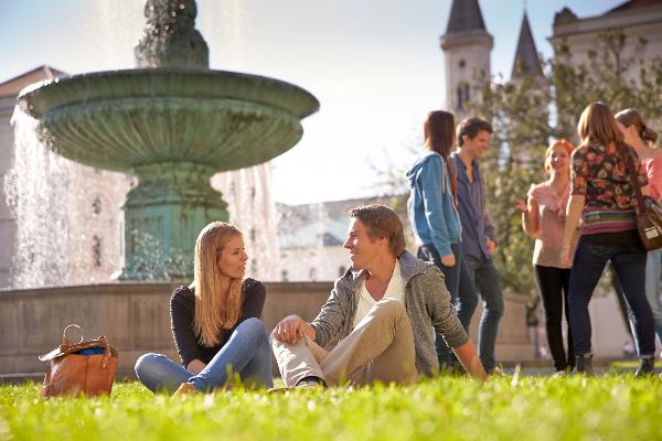 Studierende sitzem auf der Wiese vor dem Brunnen am Geschwister-Scholl-Platz.