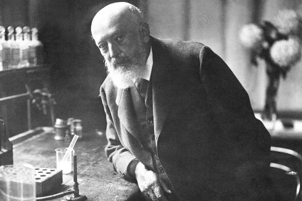 Historic black-and-white photograph of Adolf von Baeyer leaning over a laboratory table with glassware, test tubes, and a Bunsen burner.