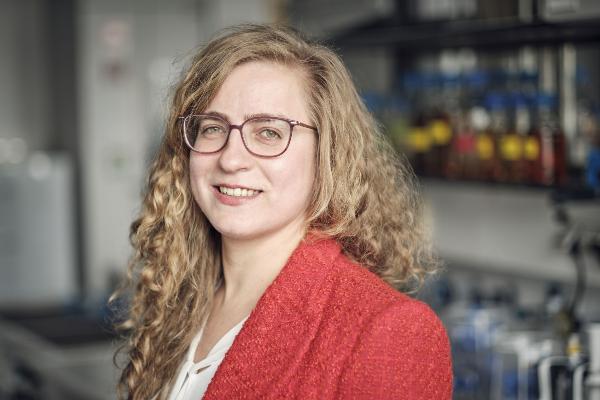 Porträt der Biochemikerin Alena Khmelinskaia. Sie trägt ein rotes Jacket und eine Brille und steht vor einem Regal mit Laborgeräten und -flaschen.