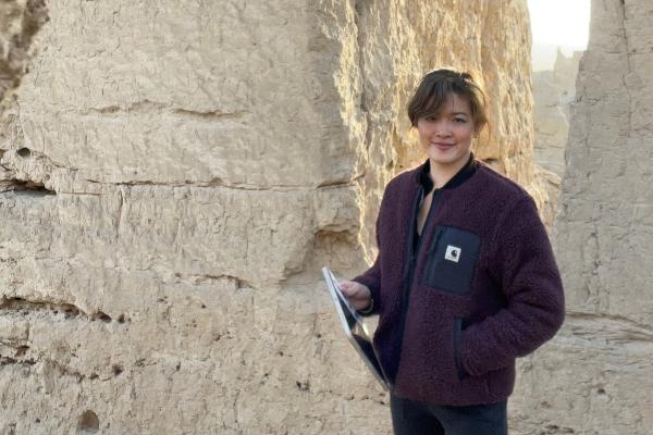 Dr. Annie Chan steht in antiker Steinruine mit Bogenfenster, trägt dunkle Jacke und hält Tablet, warmes Sonnenlicht