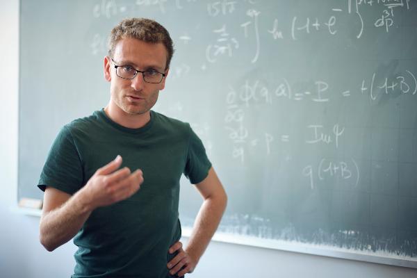 Professor Triay with glasses and in olive T-shirt with light gestures in front of a chalkboard with numerous mathematical calculations.