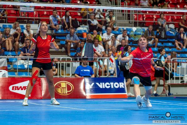 Tanja Eberl und ihre Doppel-Partnerin treten im roten Trikot in einem intensiven Badminton-Doppelspiel auf blauem Court vor Zuschauern an.