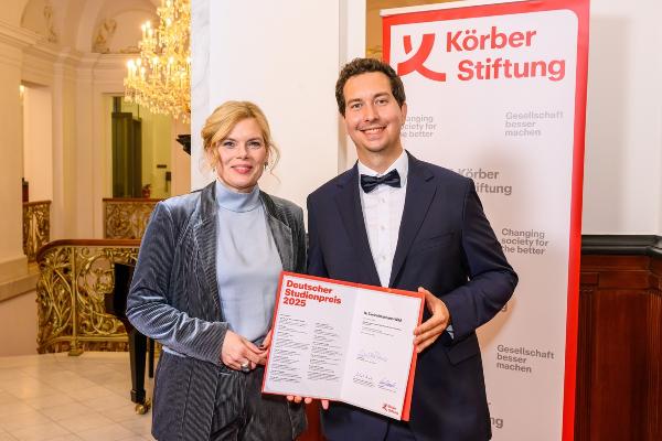 Tim Heinkelmann-Wild und President of the Bundestag Julia Klöckner pose at an award ceremony in front of a Körber Foundation banner. Heinkelmann-Wild holding a certificate.