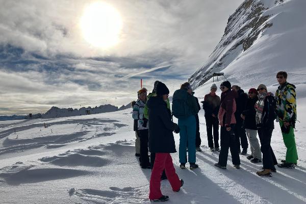 Mehrere Studierende im Winterfunktionskleidung stehen in einer Gruppe zusammen auf der Zugspitze. Im Hintergrund ist ein Lift zu sehen. Die Sonne scheint und wird vom Schnee reflektiert.