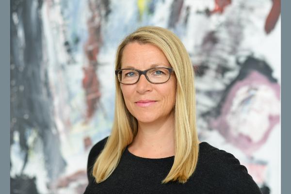Christine Bortenlänger mit blonden, langen Haaren und Brille steht vor einem abstrakten Kunstwerk. Sie trägt ein schwarzes Oberteil und schaut freundlich in die Kamera.