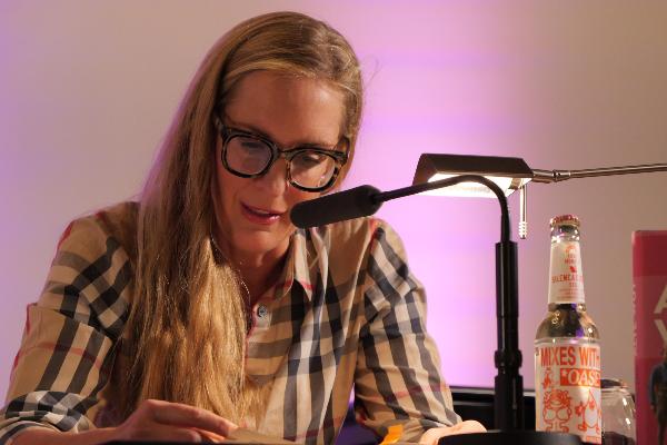 Author Caro Matzko reads from her book Alte Wut at a book reading, seated at a lectern with a microphone and reading light.