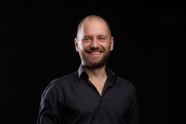 David Reichelt rasiertem Kopf und Bart, trägt ein schwarzes Hemd und eine Partitur mit einem Dirigentenstab in den Händen hält.
