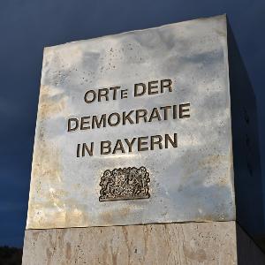 Gedenkstele „Orte der Demokratie in Bayern“ mit Bayerischem Staatswappen vor dramatisch dunklem Himmel. Das Denkmal steht symbolisch für die Demokratiegeschichte in Bayern und erinnert an bedeutende historische Orte demokratischer Entwicklung.