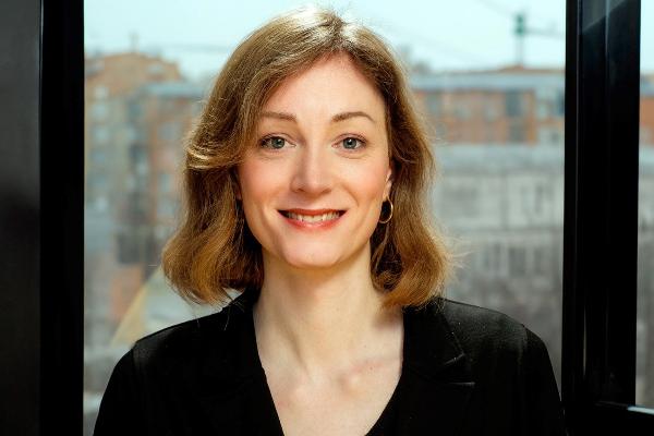Porträt einer lächelnden Frau mit kinnlangem, welligem, hellbraunem Haar, goldenen Ohrringen und schwarzer Kleidung vor einem Fenster mit Stadtblick.