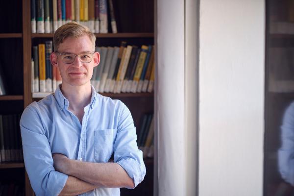 Professor Daniel Wilhelm steht vor einem Bücherregal und blickt frontal in die Kamera. Er trägt eine Brille und ein blaues Hemd.