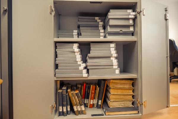 Offener Archivschrank mit grauen Archivboxen, alten Büchern und großformatigen gebundenen Bänden auf den Regalböden.