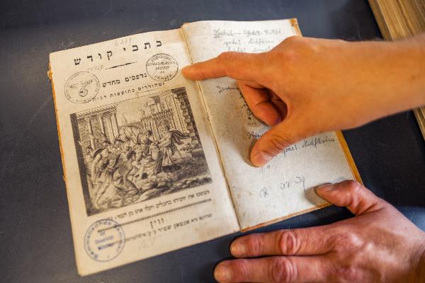 Hand zeigt auf ein altes hebräisches Buch mit Illustrationen und Stempeln, darunter ein Reichsadler und jüdische Schulstempel.