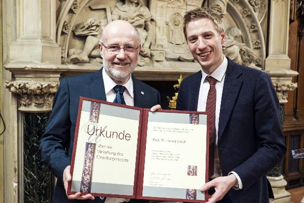 Harald Lesch und Dominik Krause in Anzügen halten gemeinsam eine Urkunde in einem historischen Raum. Die Urkunde ist vom Oberbürgermeister der Landeshauptstadt München und bescheinigt die Verleihung des Ehrenbürgerrechts an Prof. Dr. Harald Lesch. Der Raum ist mit kunstvollen Steinmetzarbeiten und einem Wappen im Hintergrund verziert.