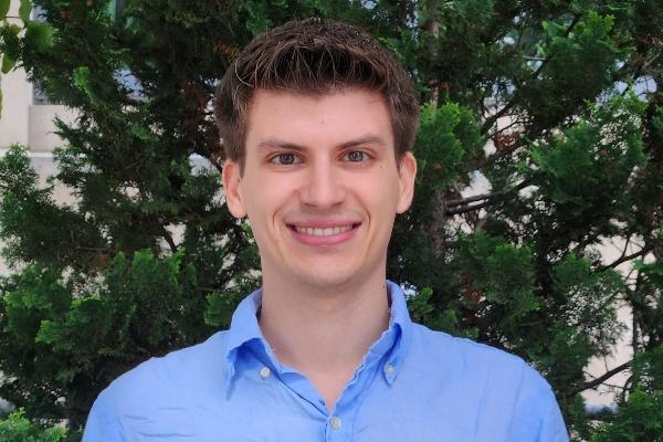 Porträt von Filip Milojevic, lächelnd, mit kurzen braunen Haaren und hellblauem Hemd vor grünen Bäumen.