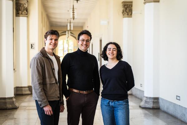 Florimon Poisson, Pablo Toussaint und Rosa Bauer are standing in a bright historic hallway of LMU´s Mainbuilding with columns, wearing modern casual clothing.
