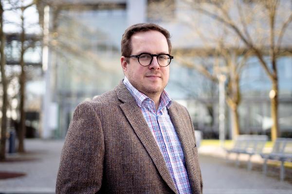 Porträt von Professor George Cutsail mit Brille, braunem Tweed-Sakko und kariertem Hemd vor modernem Glasgebäude im Freien