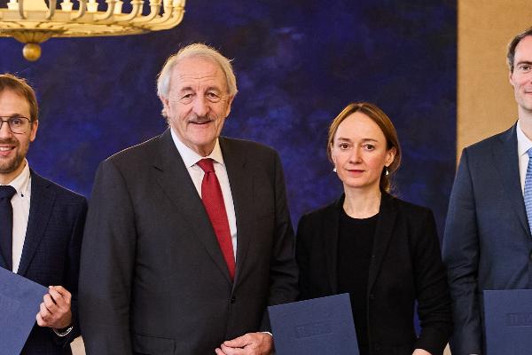 Vier Personen in formeller Kleidung halten Urkunden bei einer feierlichen Preisverleihung vor blauem Hintergrund.