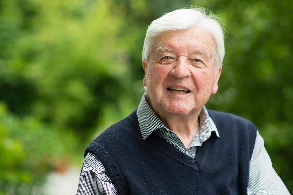 Portrait of Hans Maier with white hair looking at the camera with a slight smile. He is wearing a light patterned shirt under a dark navy sweater vest. The background is softly blurred green foliage, creating a natural and welcoming atmosphere.