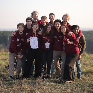 **SEO-tauglicher Alt-Text:** Gruppe junger Teilnehmer\:innen des International Physicists´ Tournament (IPT) 2025 steht lachend auf einer Wiese vor einem bewaldeten Panorama; sie tragen einheitliche bordeauxrote Hoodies mit IPT-Logo und halten stolz eine Urkunde in die Kamera.