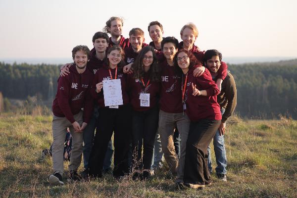 **SEO-tauglicher Alt-Text:** Gruppe junger Teilnehmer\:innen des International Physicists´ Tournament (IPT) 2025 steht lachend auf einer Wiese vor einem bewaldeten Panorama; sie tragen einheitliche bordeauxrote Hoodies mit IPT-Logo und halten stolz eine Urkunde in die Kamera.
