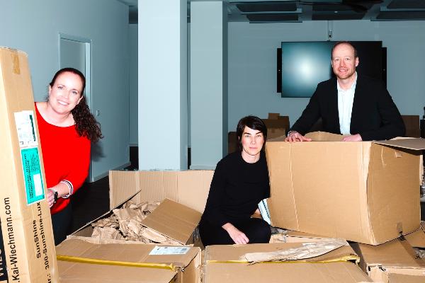 Three persons unpacking moving boxes in a modern office featuring acoustic ceiling panels, ceiling fans, and conference room technology. Office setup and workspace organization during a corporate relocation at a bright business location.