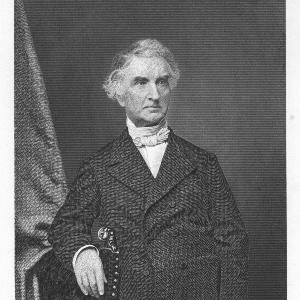 Historic engraving of the older Justus von Liebig with curly hair in a formal 19th-century suit, standing beside a chair with a draped curtain.