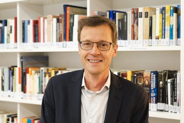 Porträt von Martin von Koppenfels mit Brille in dunklem Sakko vor gut gefüllten Bücherregalen in einer modernen Bibliothek oder Bildungseinrichtung. Professionelle Aufnahme in heller Innenraumumgebung für die Themen Bildung, Wissenschaft, Forschung und Kultur.