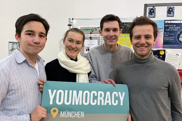 Auf dem Bild sind die vier Mitglieder des Teams von Youmocracy München zu sehen. Sie halten ein Schild mit der Aufschrift Youmocracy ins Bild.